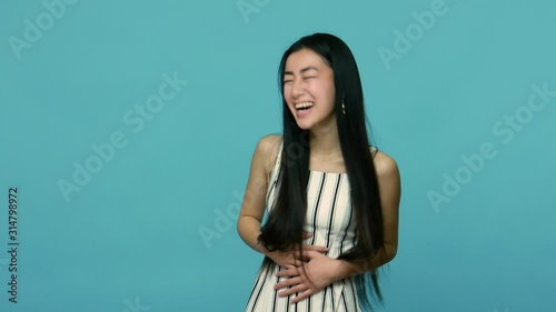 Joyful asian woman with long straight black hair in dress holding her belly and cracking up, laughing out loudly, being hysterical at something funny. indoor studio shot isolated on blue background