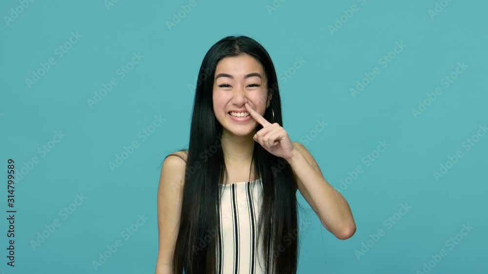 Funny crazy asian woman with long black hair showing tongue and pulling out boogers, picking her nose with stupid comical expression, making faces. indoor studio shot isolated on blue background Funny crazy asian woman with long black hair showing tongue and pulling out boogers, picking her nose with stupid comical expression, making faces. indoor studio shot isolated on blue background