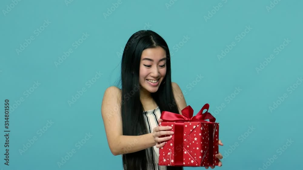 Joyful asian woman with long black hair catching wrapped present box and looking with delight and amazement at camera, rejoicing at unexpected gift. indoor studio shot isolated on blue background