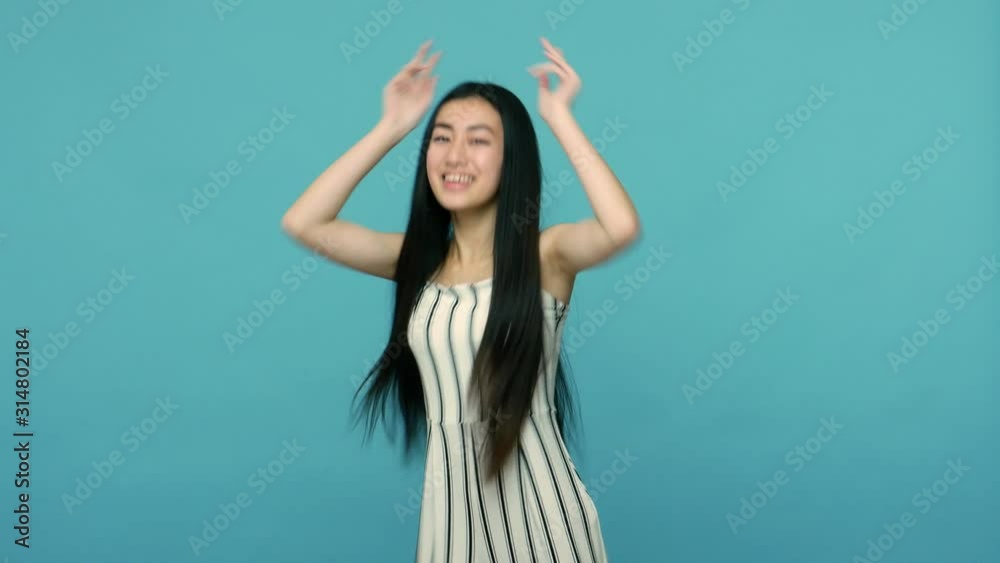 Lovely beautiful asian woman with long straight black hair and in dress dancing full of energy and joy, listening to vivid club music, quick rhythm. indoor studio shot isolated on blue background