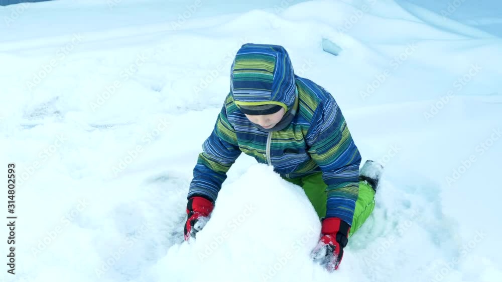 One funny child in colorful winter clothes in scarves and mittens is building tower of snow or snowman in small yard. Street entertainment for winter holidays. Snow angel One funny child in colorful winter clothes in scarves and mittens is building tower of snow or snowman in small yard. Street entertainment for winter holidays. Snow angel