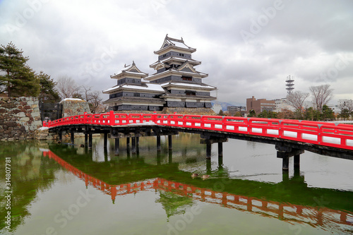 Matsumoto castle with red bridge and it’s reflection over water against cloudy sky, know as crow castle since black exterior decoration, a famous Traditional Japanese architecture in Nagano, Japan