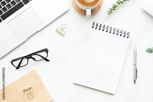 White office desk table with blank notebook, laptop computer, cup of latte coffee and supplies. Top view with copy space, flat lay.