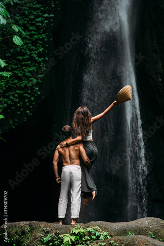 A man holds a girl in his arms at the waterfall. Couple at the waterfall, rear view. Honeymoon trip. Happy couple on vacation in Bali. A couple in love travels the world. Vacation in Asia, Indonesia.