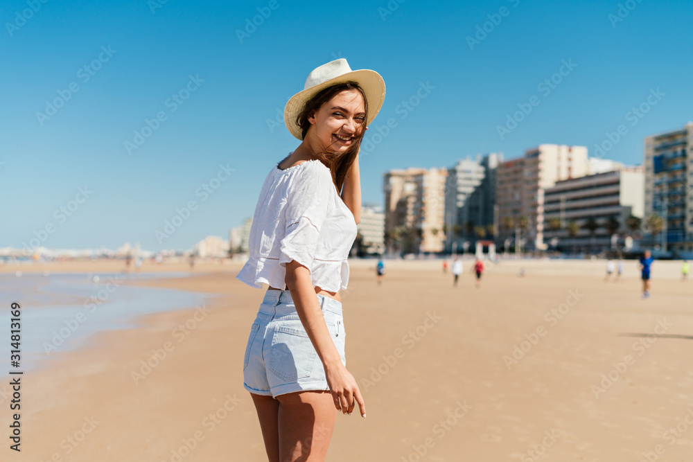 smiling girl is standing on the beach sand looking over her shoulder, behind the building, people are walking in the distance smiling girl is standing on the beach sand looking over her shoulder, behind the building, people are walking in the distance