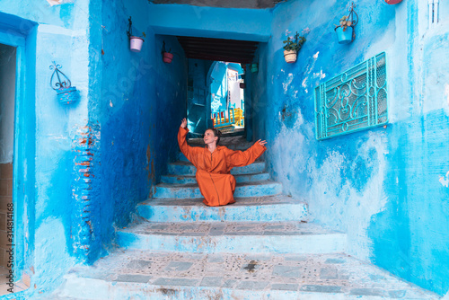 girl in a bright orange African dress sits on the steps of the narrow street of the blue city of Morocco © nelen.ru