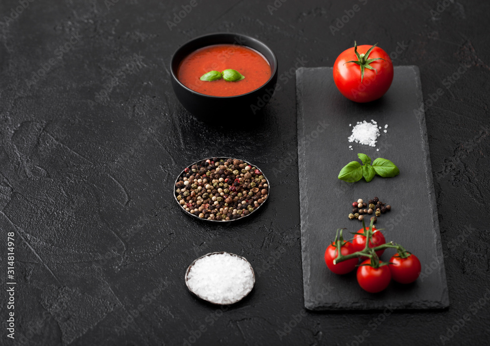 Black bowl plate of creamy tomato soup on black table background with stone chopping board and raw tomatoes, pepper and salt. Black bowl plate of creamy tomato soup on black table background with stone chopping board and raw tomatoes, pepper and salt.