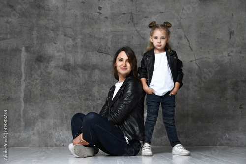 Positive young woman and cute little girl in similar rock style clothing and black leather jackets posing over grey background