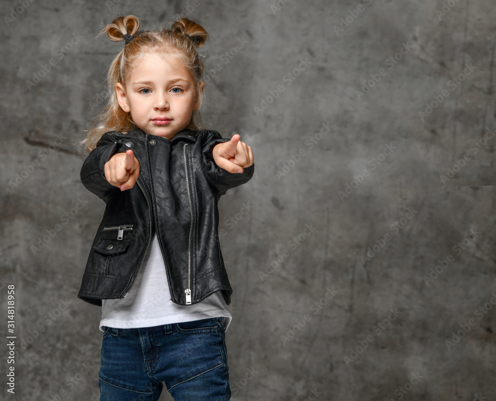 Little cute blond girl in stylish rock style black leather jacket, jeans and sneakers standing and pointing at camera with fingers Little cute blond girl in stylish rock style black leather jacket, jeans and sneakers standing and pointing at camera with fingers