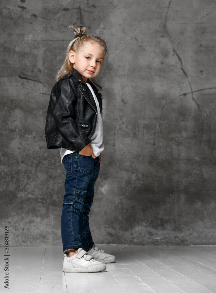 Little smiling blond girl in stylish rock style black leather jacket, blue jeans and white sneakers standing with hands in pockets