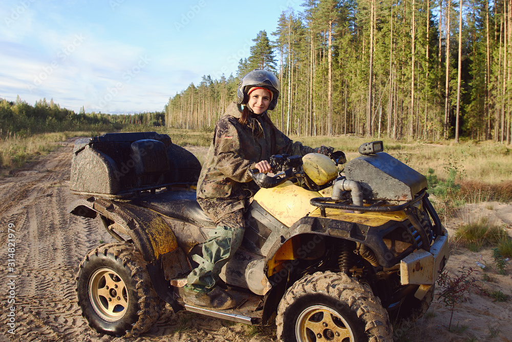 Fototapeta premium Young beautiful tanned woman dressed in special clothes under disguise wears helmet with glasses rides powerful Quad bike with large studded wheels, girl smiles, looks happy. Against background forest