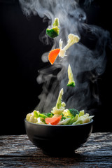 The steam from the vegetables carrot broccoli cauliflower on black bowl , a steaming. Boiled hot Healthy food on table on black background,hot food and healthy meal concept
