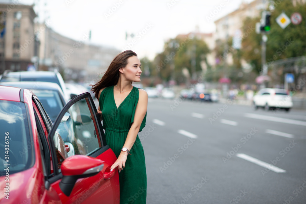Fashion model wearing green overall posing outdoor next to her red car on the road. Young beautiful brunette caucasian woman driver. Beautiful girl, urban portrait. Fashion model wearing green overall posing outdoor next to her red car on the road. Young beautiful brunette caucasian woman driver. Beautiful girl, urban portrait.