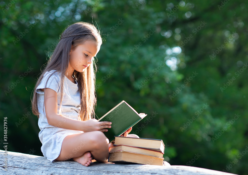 Obraz premium Charming little girl with long brown hair reads book outdoor sitting on tree in summer park or in a forest glade. Child booklover