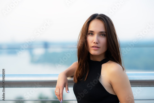 Fashion model wearing black elegant dress posing outdoor at the bank of Dniper river in Kyiv, Ukraine. Young beautiful brunette caucasian woman walking summer streets. Beautiful girl, urban portrait.