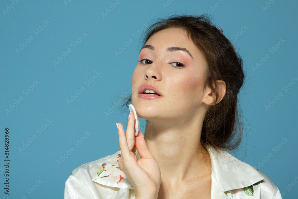 Cheerful attractive teen girl clean her face with cotton disk, isolated over blue background. Portrait of young beautiful woman during morning procedures Cheerful attractive teen girl clean her face with cotton disk, isolated over blue background. Portrait of young beautiful woman during morning procedures