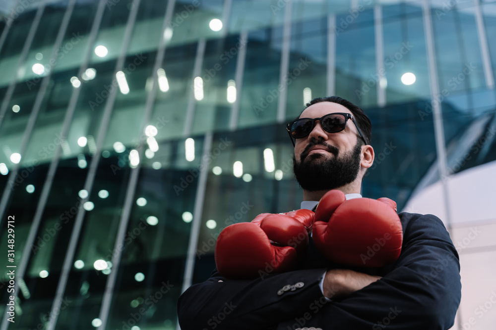 Businessman smiling with sunglasses and red boxing gloves hanging from his neck with his arms crossed in front of a building Businessman smiling with sunglasses and red boxing gloves hanging from his neck with his arms crossed in front of a building
