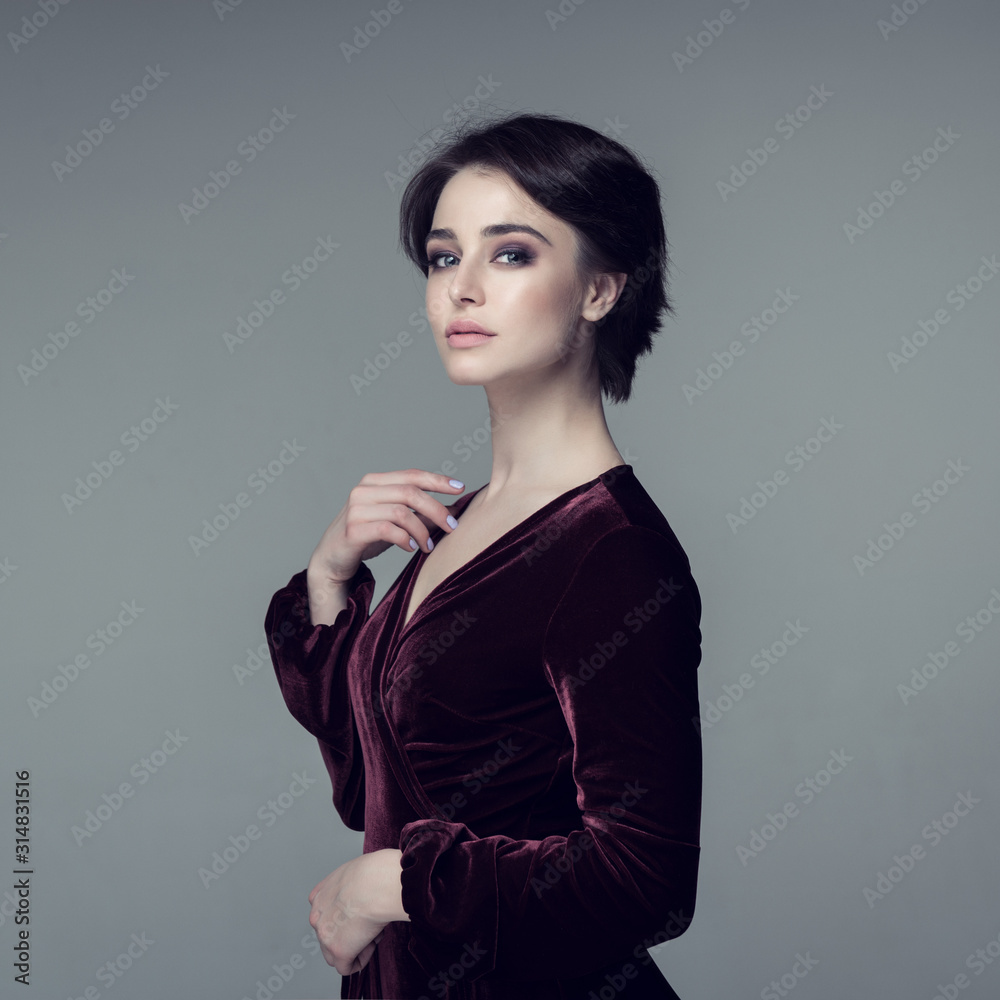 Beauty female portrait. Young attractive woman posing at studio over gray background. Beautiful model with perfect make-up dressed evening burgundy dress looking at camera. Beauty female portrait. Young attractive woman posing at studio over gray background. Beautiful model with perfect make-up dressed evening burgundy dress looking at camera.