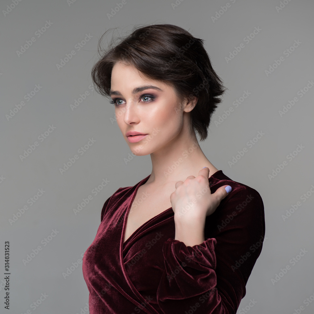 Fototapeta premium Beauty female portrait. Young attractive woman posing at studio over gray background. Beautiful model with perfect make-up dressed evening burgundy dress looking at camera.