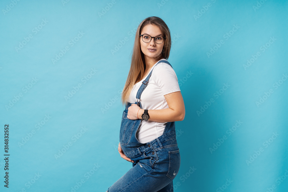 Smiling pregnant woman wearing glasses caressing her belly over blue background Smiling pregnant woman wearing glasses caressing her belly over blue background