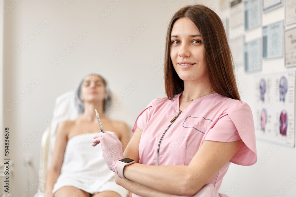Close up portrait of cosmetologist finishes facial rejuvenation procedure for her client, posing near her patient who relaxes on couch, looking directly at camera. Skin care, cosmetology concept. Close up portrait of cosmetologist finishes facial rejuvenation procedure for her client, posing near her patient who relaxes on couch, looking directly at camera. Skin care, cosmetology concept.