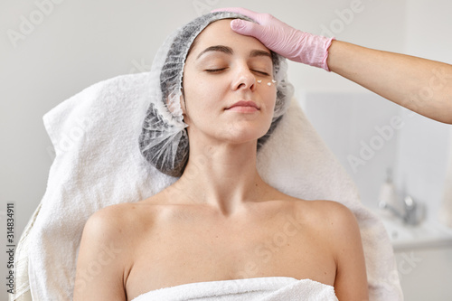 Close up portrait of young woman spends time in beauty parlor, being readfy for cleansing massage. Proffesional Cosmetologist examines attractive female dresses medical cap, posing with closed eyes.