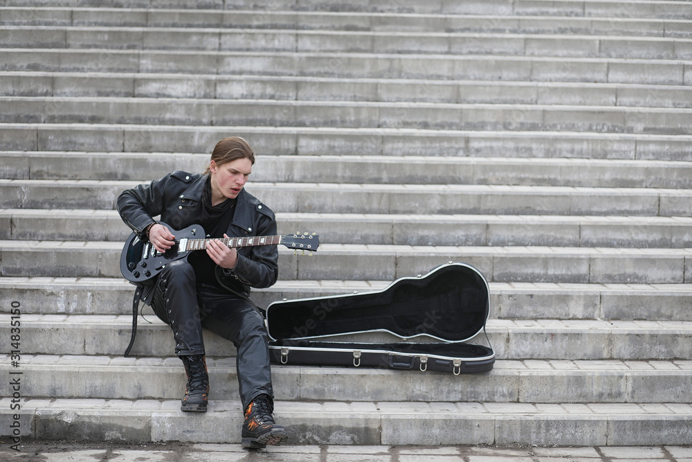 Rock guitarist on the steps. A musician with a bass guitar in a leather suit. Metalist with a guitar on the background of industrial steps.