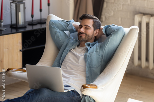 Peaceful young man daydreaming in comfortable armchair, holding computer. photo