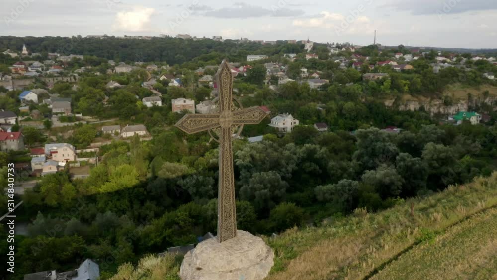 The beautifully metal cross on a concrete mound and situated on the edge of a mountain, overlooking a peaceful city of Ukraine with the Kamianets-Podilskyi castle in the distance - Aerial shot The beautifully metal cross on a concrete mound and situated on the edge of a mountain, overlooking a peaceful city of Ukraine with the Kamianets-Podilskyi castle in the distance - Aerial shot