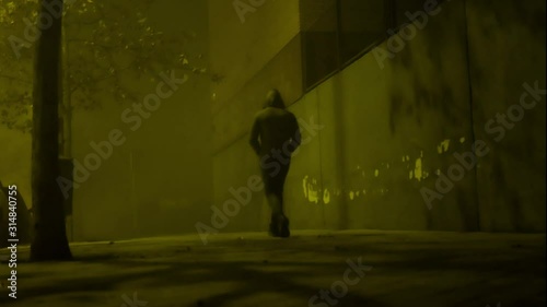 A shot at ground level of a hooded man walking away near a wall in a foggy night
