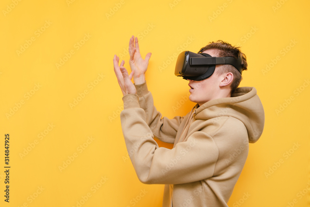 Portrait of surprised guy in VR helmet stands on yellow background and looks at his hands. Young man in casual clothes uses a virtual reality helmet. Isolated. Portrait of surprised guy in VR helmet stands on yellow background and looks at his hands. Young man in casual clothes uses a virtual reality helmet. Isolated.