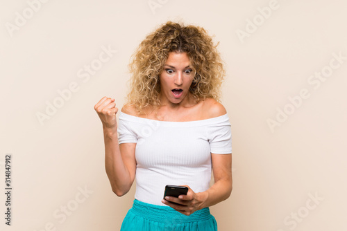 Young blonde woman with curly hair over isolated background surprised and sending a message