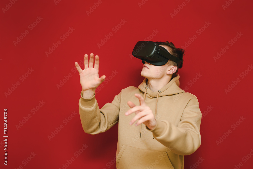Portrait of a young man in casual clothes testing a VR helmet on a red background, looking away and touching his hands. Guy in the virtual reality helmet is isolated on a yellow background. Portrait of a young man in casual clothes testing a VR helmet on a red background, looking away and touching his hands. Guy in the virtual reality helmet is isolated on a yellow background.