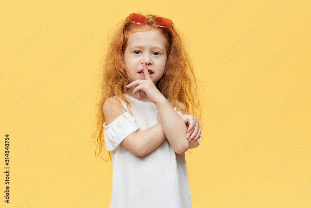 Signs, gestures and symbols concept. Charming adorable Caucasian girl asking for silence, making shush gesture with fore finger at her mouth posing isolated against blank yellow wall background Signs, gestures and symbols concept. Charming adorable Caucasian girl asking for silence, making shush gesture with fore finger at her mouth posing isolated against blank yellow wall background