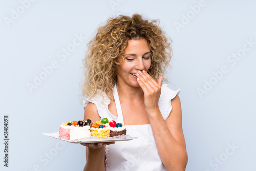 Young blonde woman with curly hair holding lots of different mini cakes smiling a lot