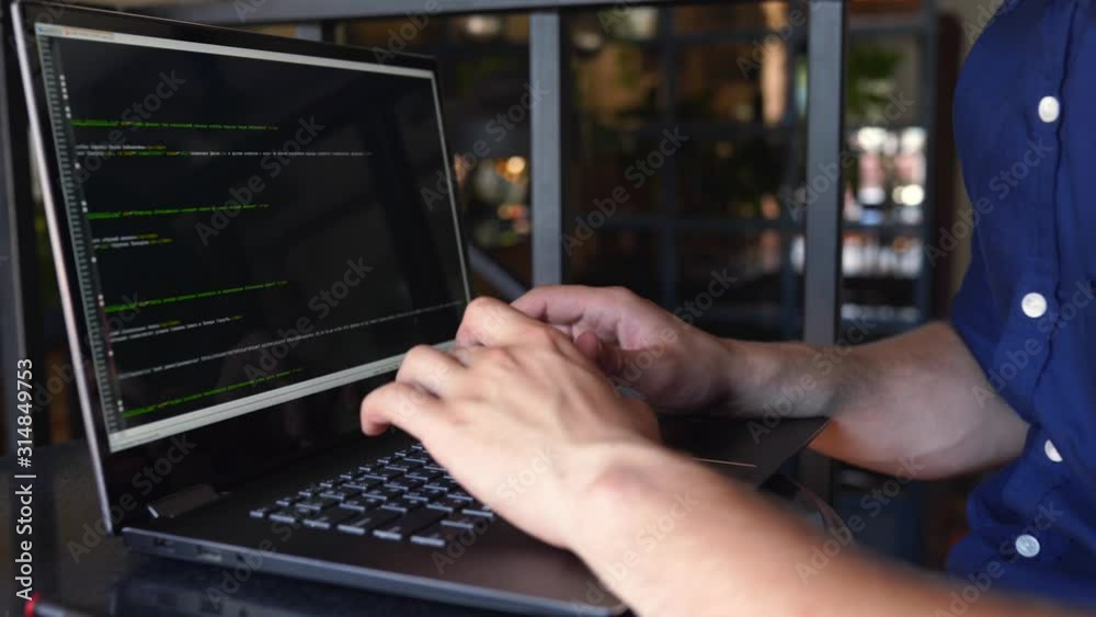 Back view over the shoulder shot of developer programmer with laptop. Program code and script data on the screen. Young freelancer in glasses working on project in cafe.