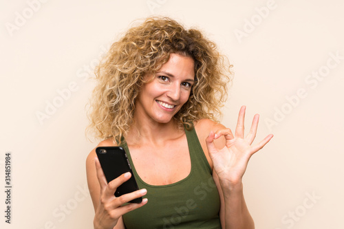 Young blonde woman with curly hair using mobile phone showing ok sign with fingers