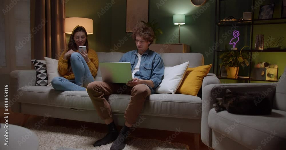 Cheerful oung Caucasian guy and girl talking and laughing while sitting on the sofa and using smartphone and laptop computer. Inside. At night. Cheerful oung Caucasian guy and girl talking and laughing while sitting on the sofa and using smartphone and laptop computer. Inside. At night.
