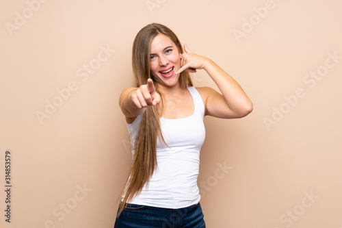 Teenager blonde girl over isolated background making phone gesture and pointing front