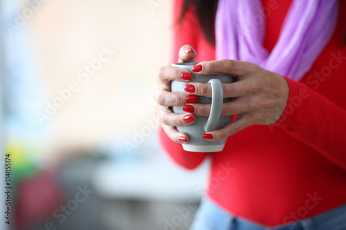 Focus on hardworking female arms holding green cup of hot beverage. Trendy woman in fashionable clothes standing in comfortable workplace studio. Fashioner workshop concept. Blurred background