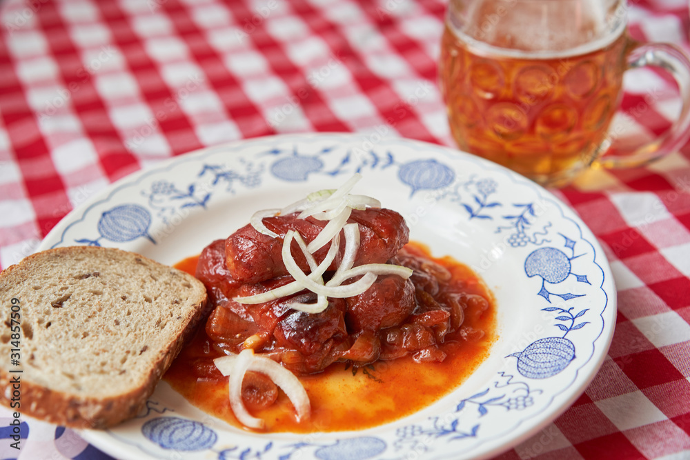 Czech food specialty, roasted small sausages in beer sauce made from dark beer, tomato sauce, chily, garlic and onion served on rustic plate with glass of lager beer and bread, typical czech pub snack Czech food specialty, roasted small sausages in beer sauce made from dark beer, tomato sauce, chily, garlic and onion served on rustic plate with glass of lager beer and bread, typical czech pub snack