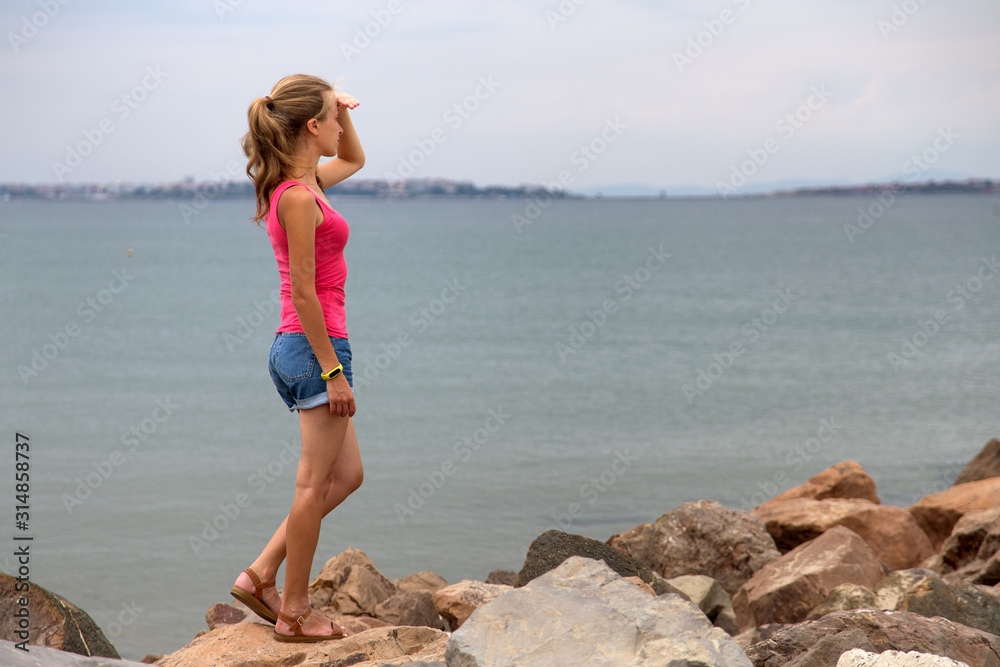 Young woman in summer clothes standing on big boulders on sea shore looking on horizon.