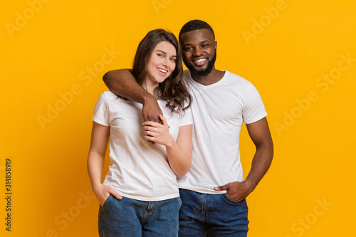 Happy multiracial couple hugging and posing over yellow background