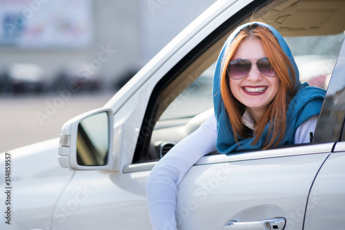 Young fashionable smiling woman driver looking out the window behind car steering wheel.