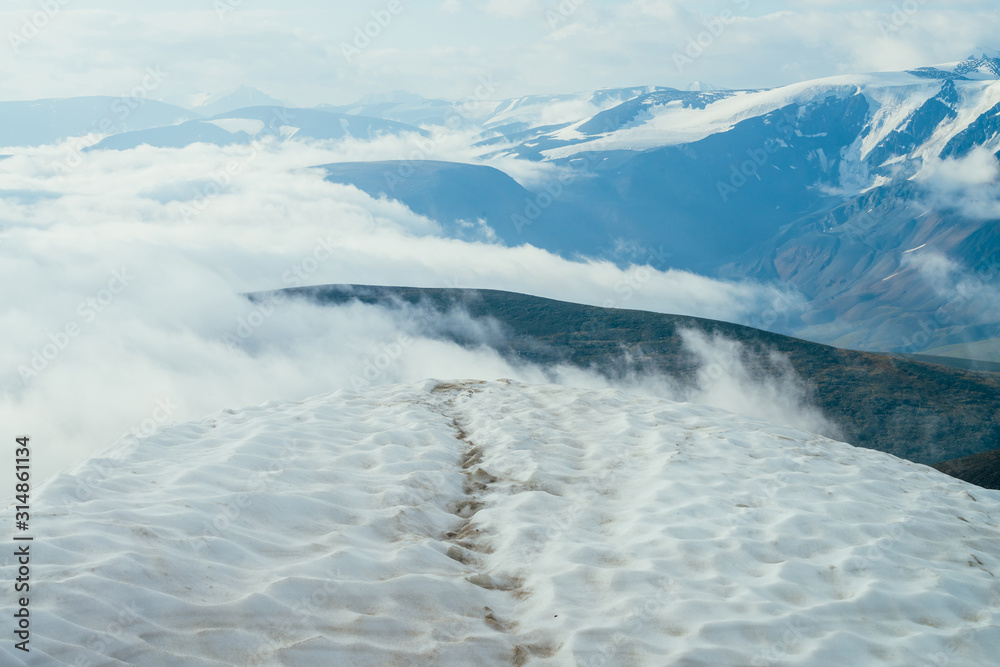 Footprints in snow on mountain top above thick clouds with view to giant mountains and glaciers. Snow on mountain peak among thick low clouds. Atmospheric alpine landscape. Wonderful highland scenery.