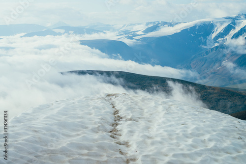 Footprints in snow on mountain top above thick clouds with view to giant mountains and glaciers. Snow on mountain peak among thick low clouds. Atmospheric alpine landscape. Wonderful highland scenery.