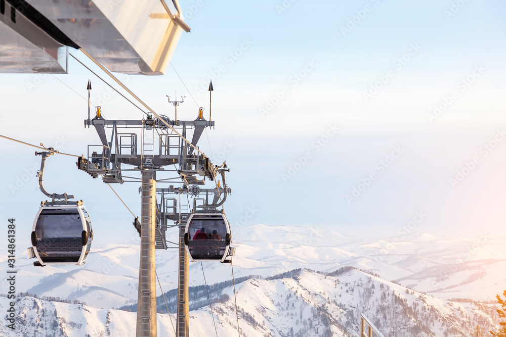 Obraz premium Cabin of a gondola cableway suspended on a rope where sits people with skis and snowboards high in the Altai mountains with snow and blue sky on winter sunset. Ski resorts and snowboarding.