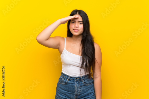 Young teenager Asian girl over isolated yellow background looking far away with hand to look something