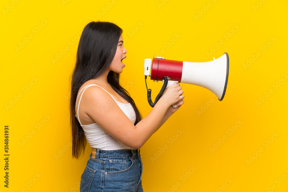 Young teenager Asian girl over isolated yellow background shouting through a megaphone Young teenager Asian girl over isolated yellow background shouting through a megaphone