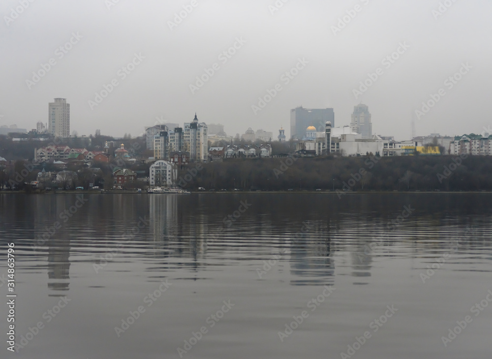 Cloudy winter day in the city. Houses, trees and bridges in the city are reflected in the water of the river on a cloudy day at the beginning of winter.
